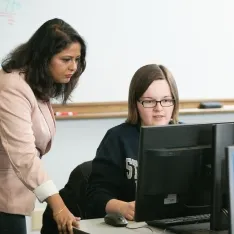 A woman helping a student at a computer in a classroom.